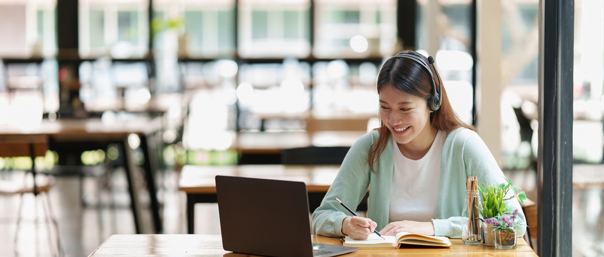 Young female wearing headset working on laptop while taking notes