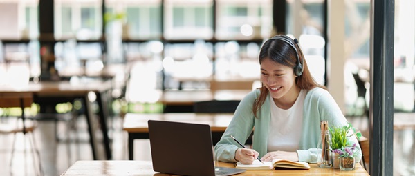 Young female wearing headset working on laptop while taking notes