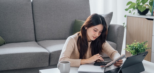 Young Asian female sitting on floor at coffee table while working on her laptop and calculator
