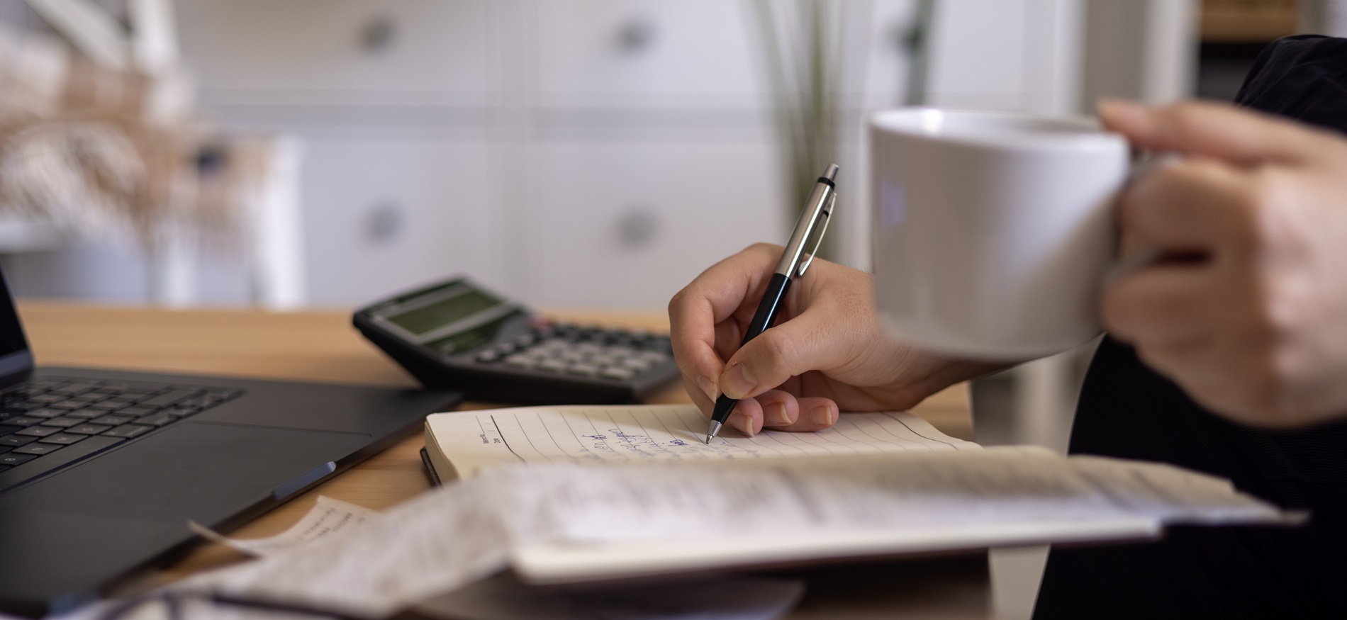 hand holding a cup of coffee while making notes and working on her laptop and calculator