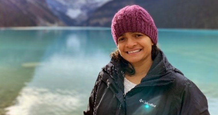 Woman in winter hat smiles in front of lake and mountain.