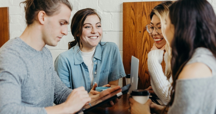 group of four people smiling and talking while holding coffees and electronic devices