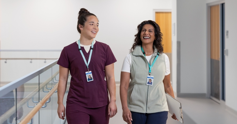 A pair of smiling female health-care workers walking together down the hall of a hospital.