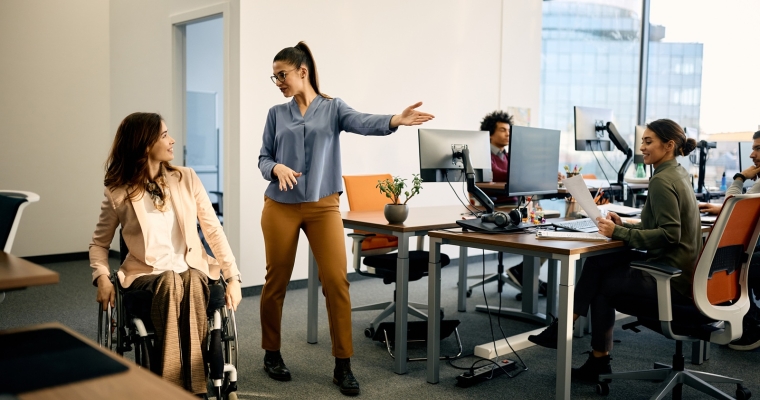 Happy businesswoman talks to her wheelchair-bound colleague while leading her through the office