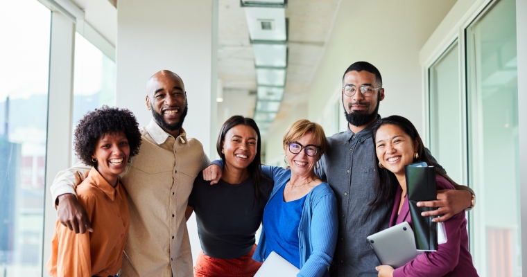 Portrait of a group of six diverse businesspeople smiling while standing arm in arm together in an office corridor.
