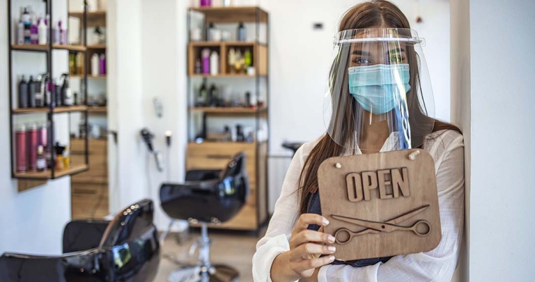 woman wearing protective mask and face shield holding open sign in front of barber shop
