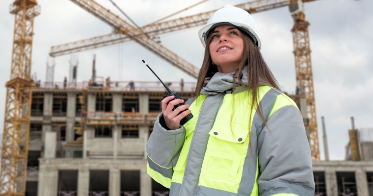 young woman in hard hat and reflective safety jacket holding a walkie talkie at a construction site