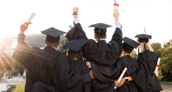 Back view of five new graduates wearing graduation caps and gowns holding their new degrees up in the air and looking out over a sunset landscape.