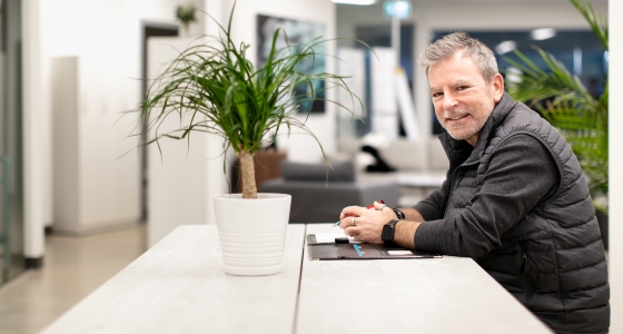 smiling man sitting at a desk in an open concept office with green plants nearby