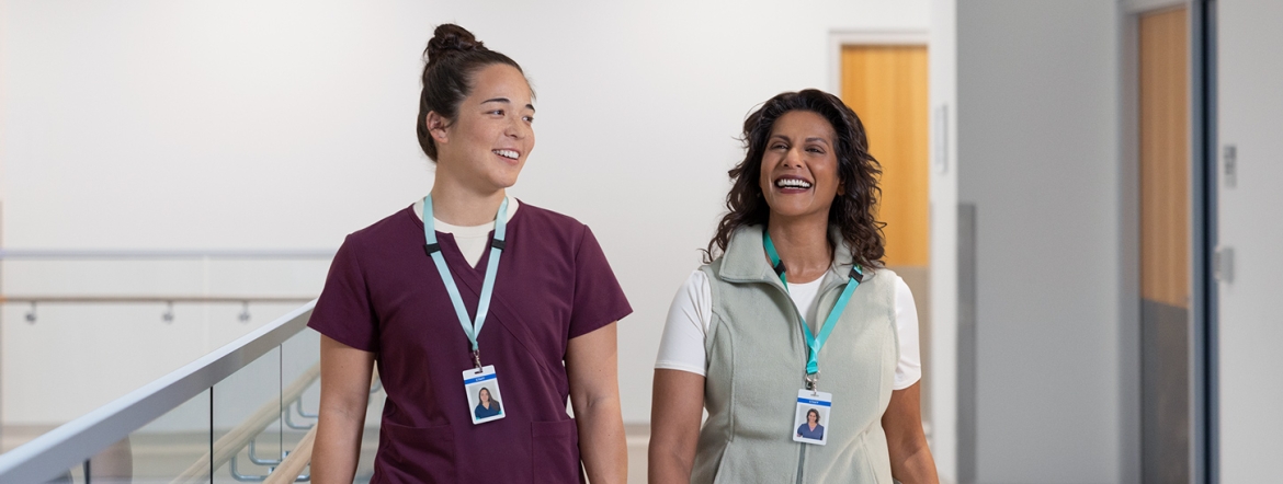A pair of smiling female health-care workers walking together down the hall of a hospital.