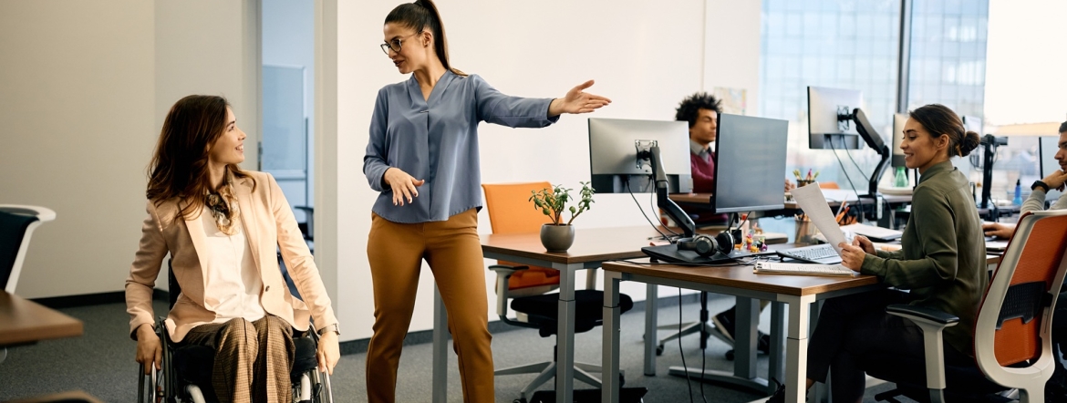 Happy businesswoman talks to her wheelchair-bound colleague while leading her through the office