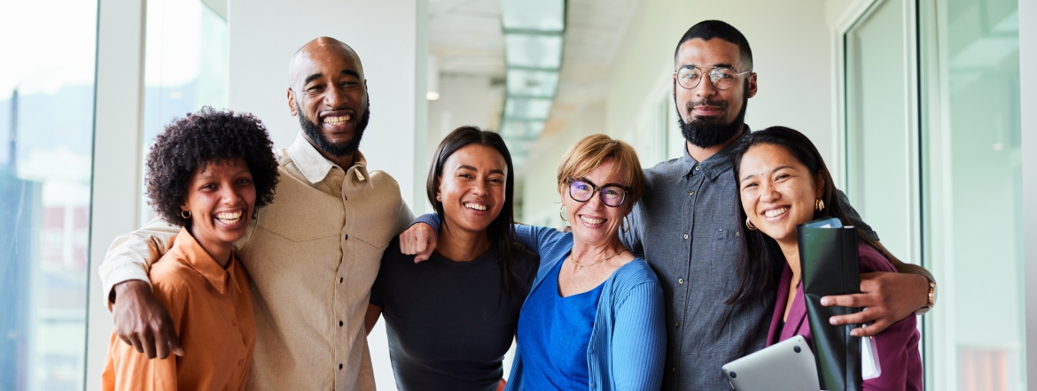 Portrait of a group of six diverse businesspeople smiling while standing arm in arm together in an office corridor.