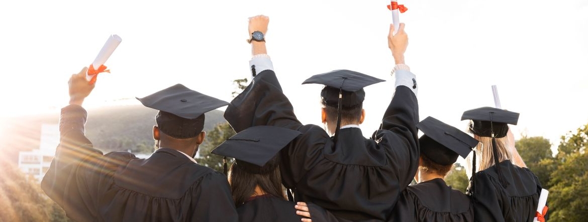 Back view of five new graduates wearing graduation caps and gowns holding their new degrees up in the air and looking out over a sunset landscape.