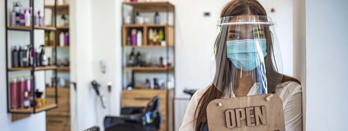 woman wearing protective mask and face shield holding open sign in front of barber shop