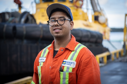 Hamza standing in front of a tugboat wearing orange work overalls
