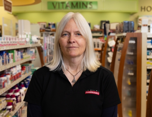 Tish standing in a retail pharmacy wearing black work shirt with red logo.