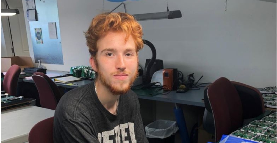Lucas faces camera while seated in front of technical equipment at his new job.
