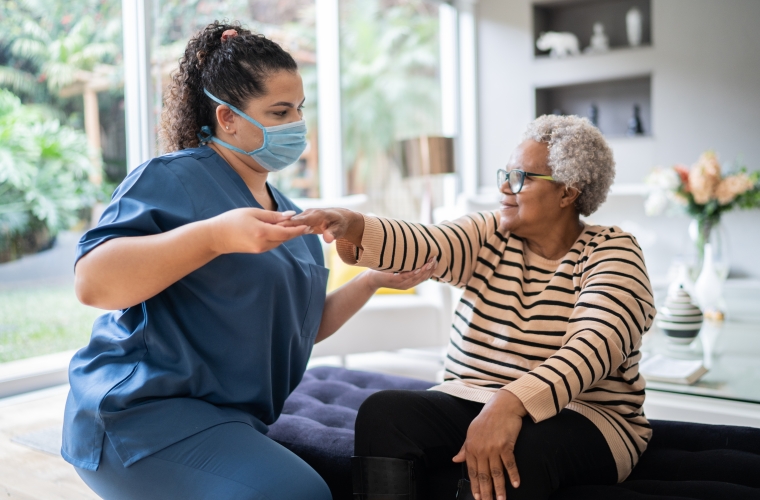 Female physical therapist wearing scrubs and a face mask supporting a senior woman doing an arm exercise at home.