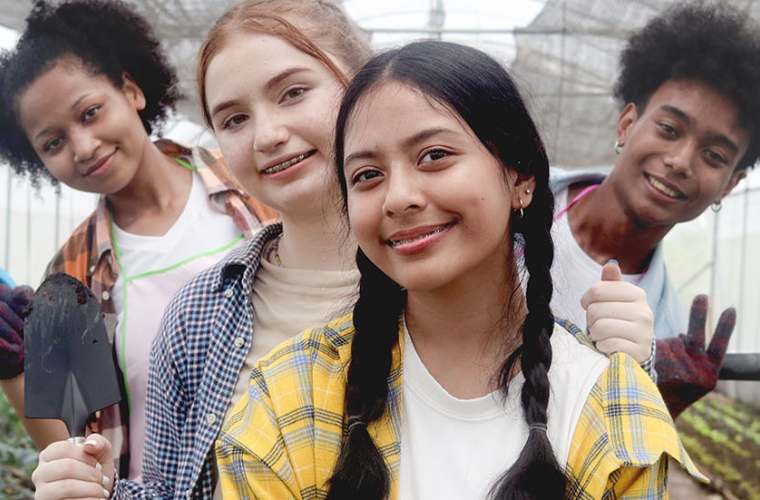 A diverse group of young people holding gardening tools and smiling in a commercial greenhouse