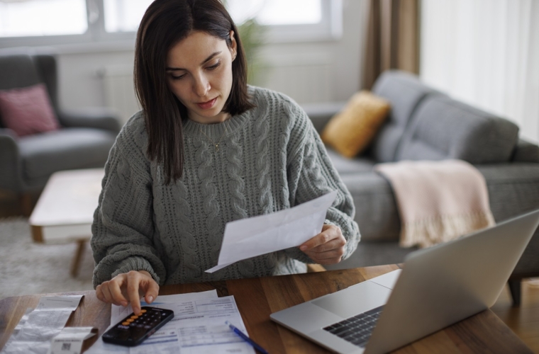 Caucasian female with dark hair reading document while using a calculator at her computer desk