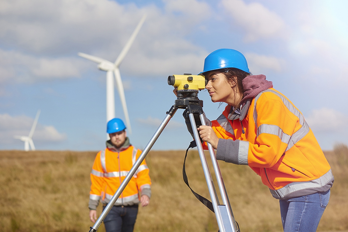 woman with hardhat using estimator with a man and windmill in background