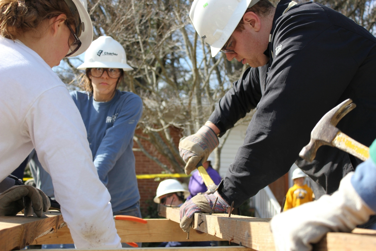 group of people wearing hard hats, safety glasses and gloves using tools to work on wooden frame outdoors