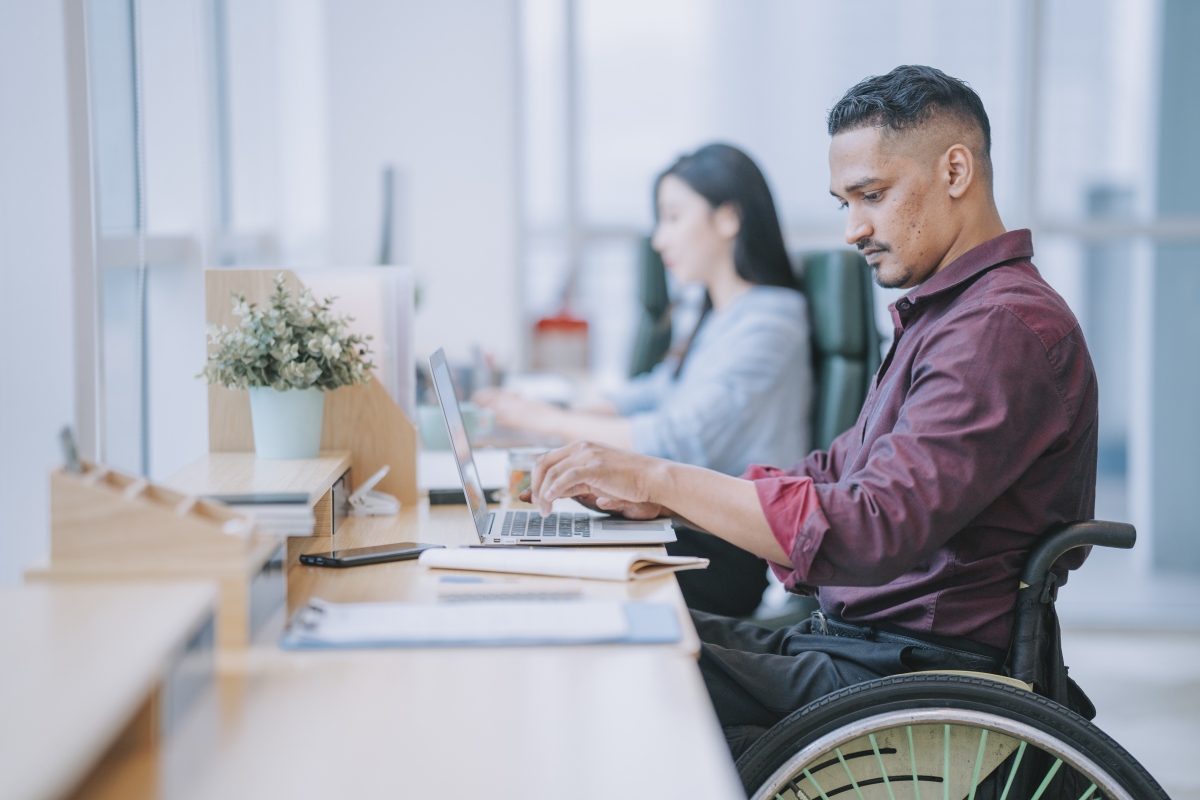 Asian Indian male worker in wheelchair concentrating working in office beside his colleague