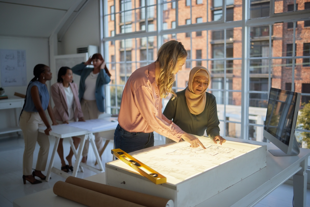 Two female coworkers reviewing blueprints on a lighted desk in office 
