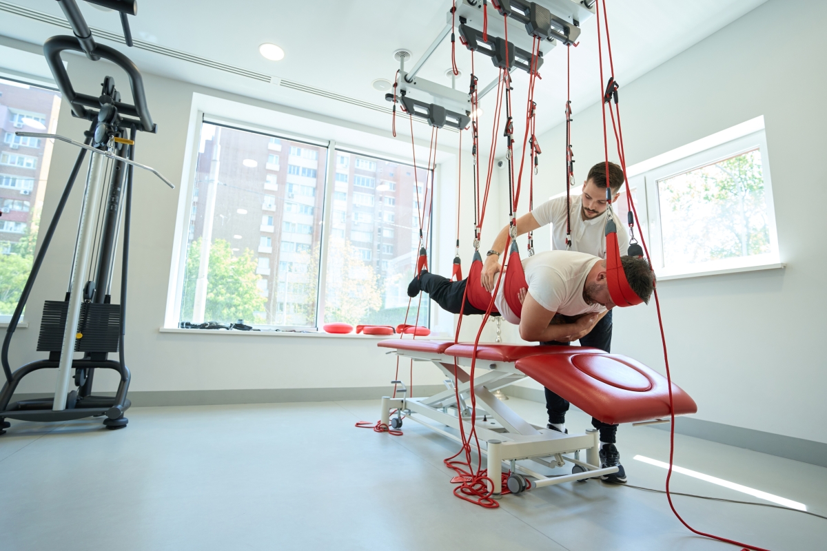Patient working out in prone position on suspension trainer during gym workout with kinesiotherapist