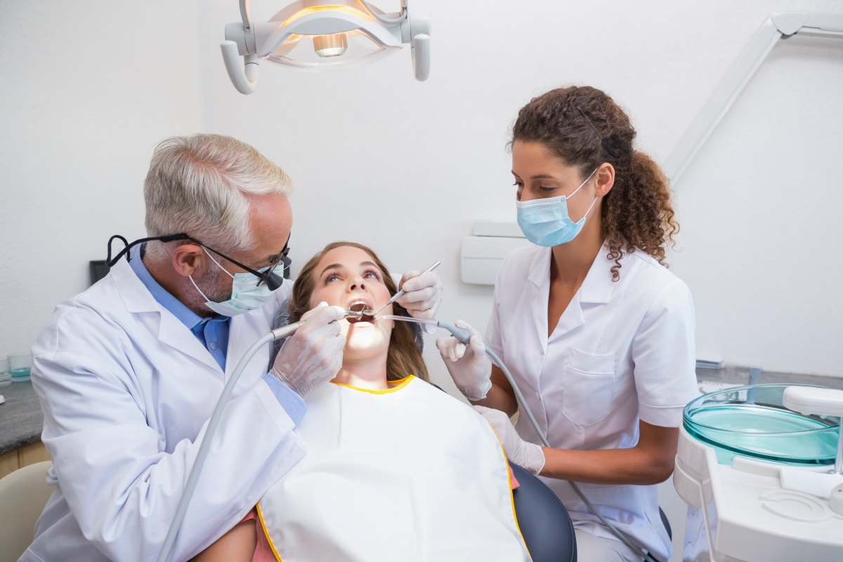 Mature male dentist examining a patients teeth in the dentists chair with assistant 
