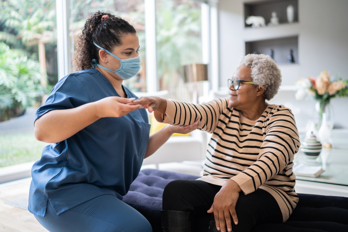 Female physical therapist wearing scrubs and a face mask supporting a senior woman doing an arm exercise at home.