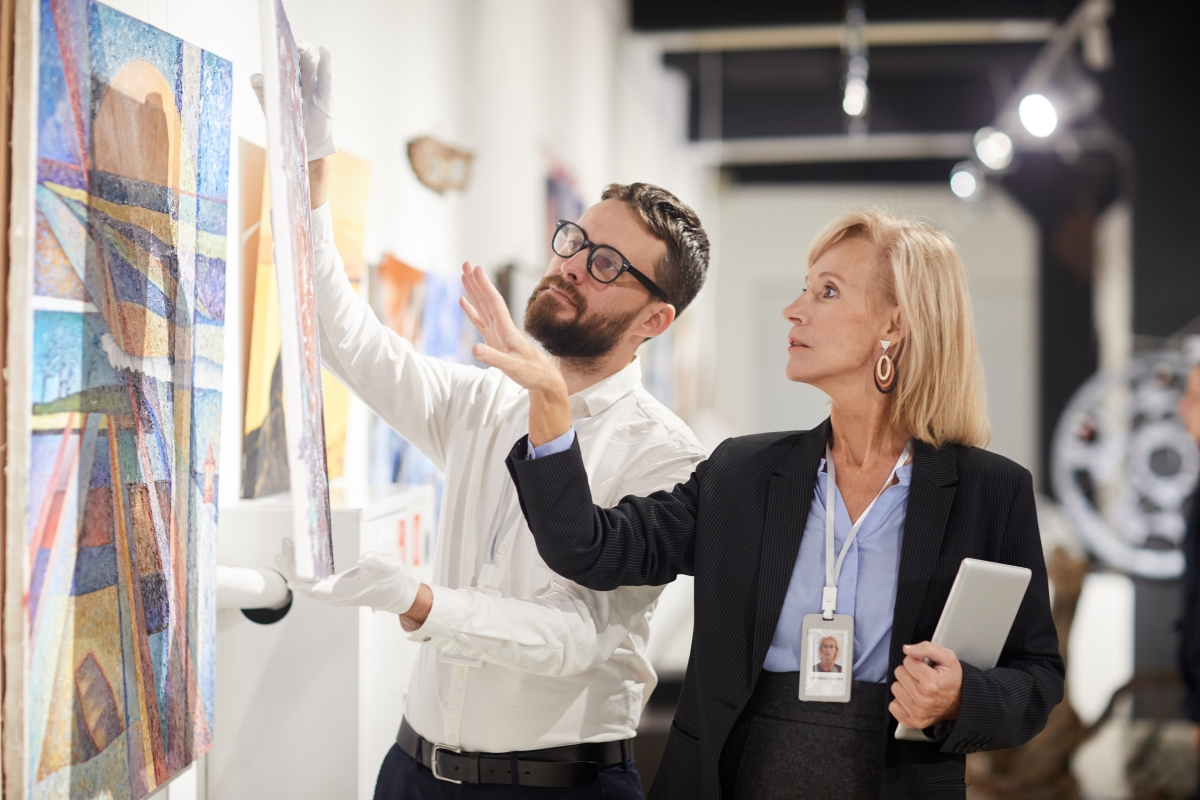 Female art gallery manager oversees her male staff hanging a painting on the wall.