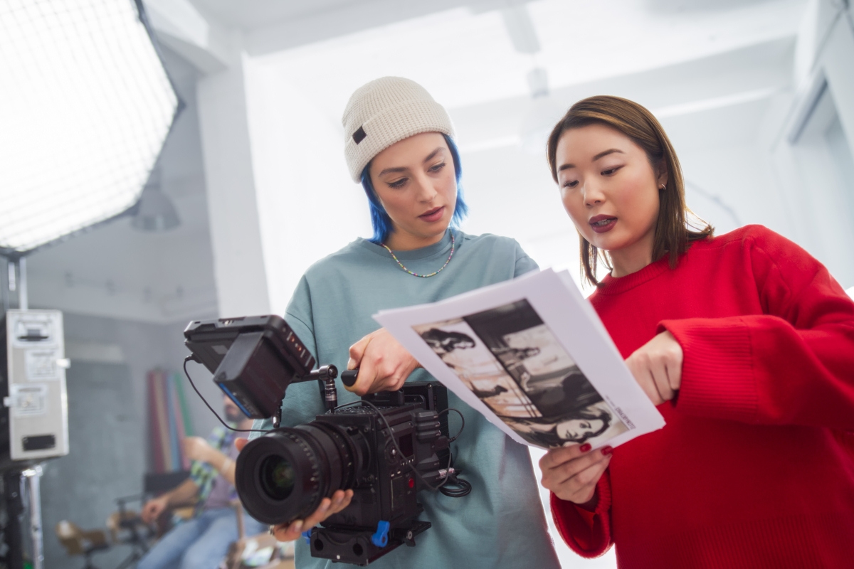 Asian woman in a red sweater and a caucasian woman with blue hair reviewing storyboard over a camera. 