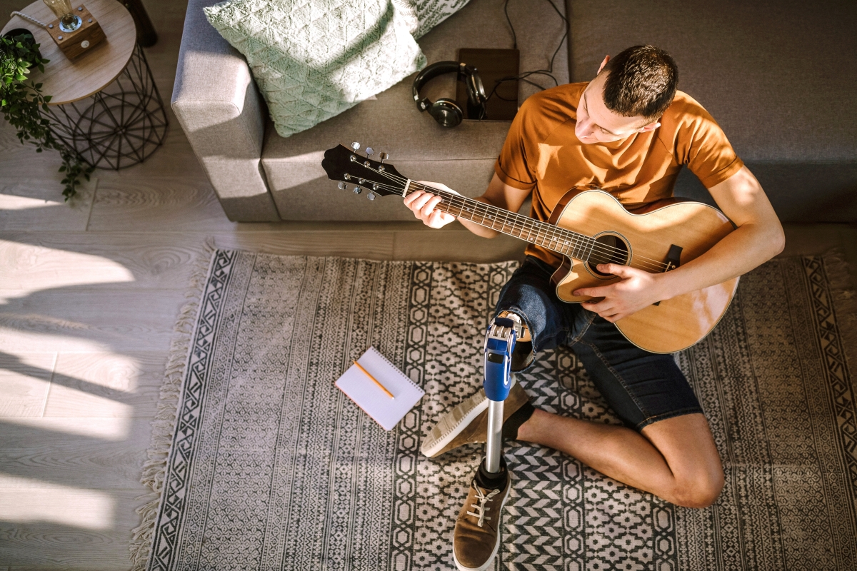 Young man with prosthetic leg sitting on floor at home and playing acoustic guitar