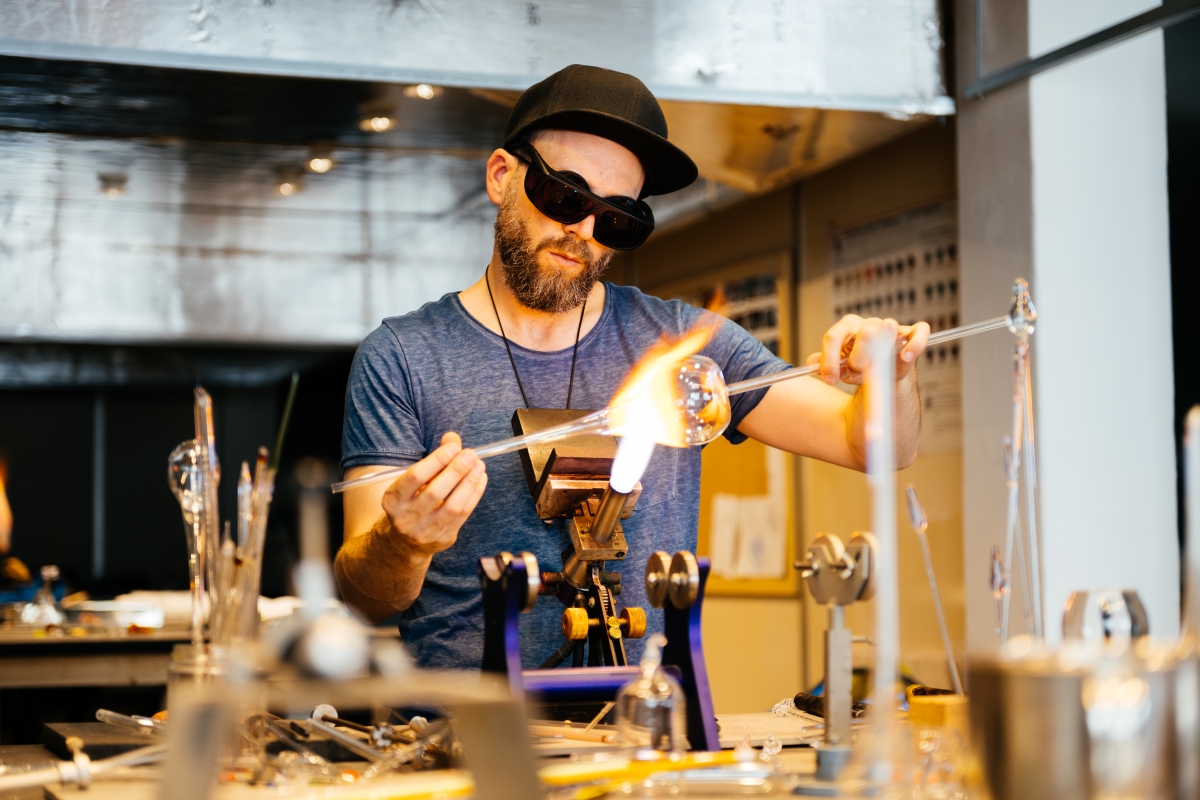 Male glassblowing artisan heating and manipulating a piece of glass with a torch and long glassblowing rod.