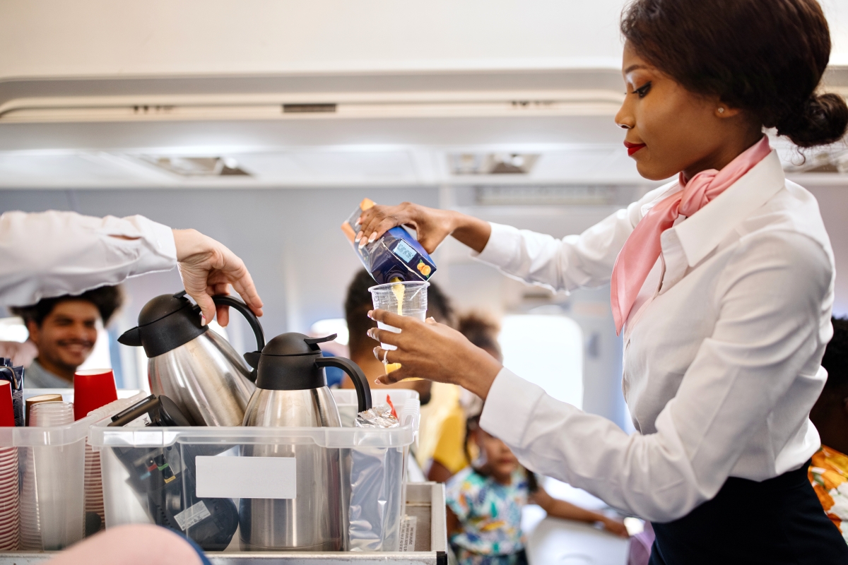 Black flight attendant pushing service cart and serving customer on airplane during flight.