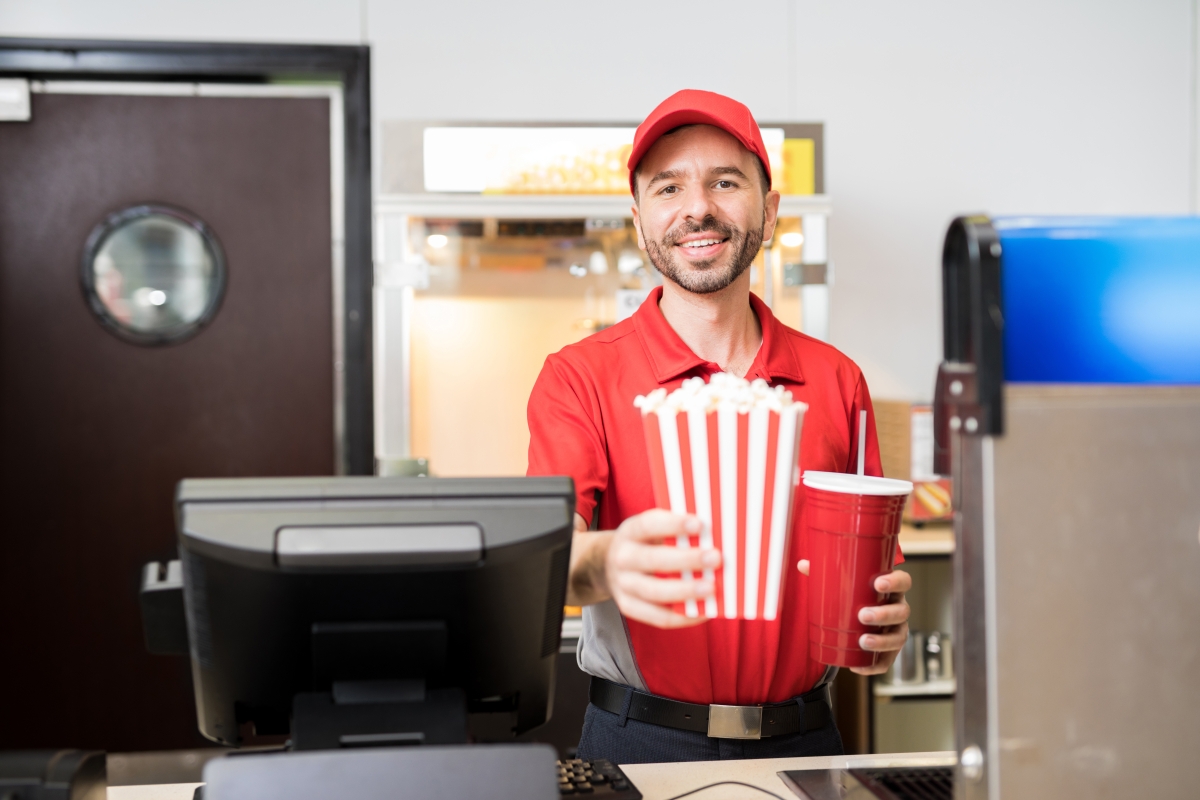 Male worker in uniform selling popcorn and soda at the concession stand in a movie theater.