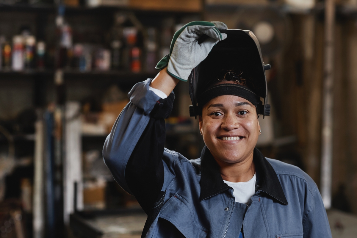 Young female welder in metal workshop holding her helmet open.
