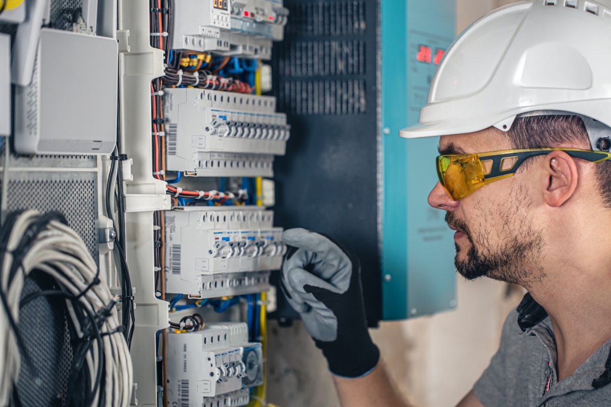 An electrical male technician wearing protective gear working on a switchboard with fuses.