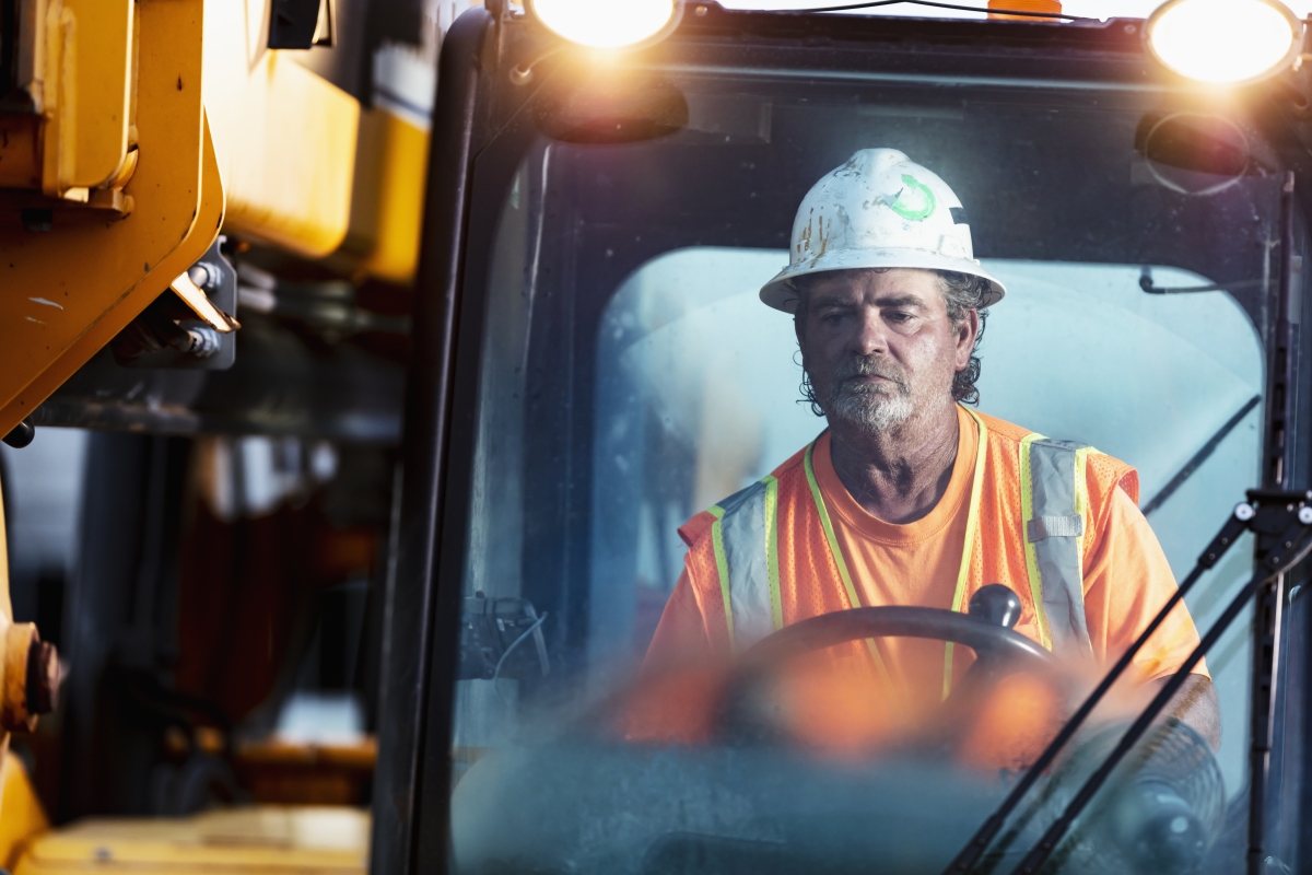 Mature male worker wearing safety gear operating heavy machinery at job site.