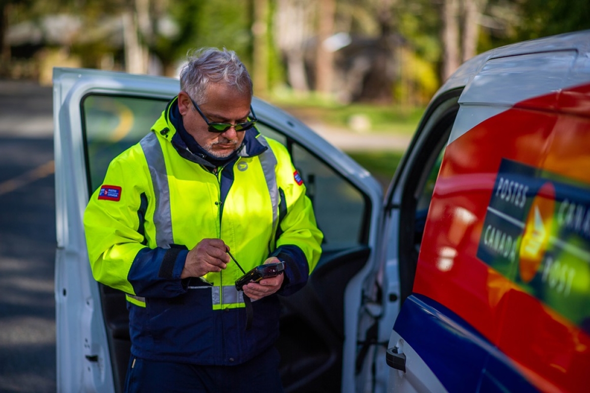 Mature male postal carrier in reflective gear by side of postal van checking phone for delivery instructions.