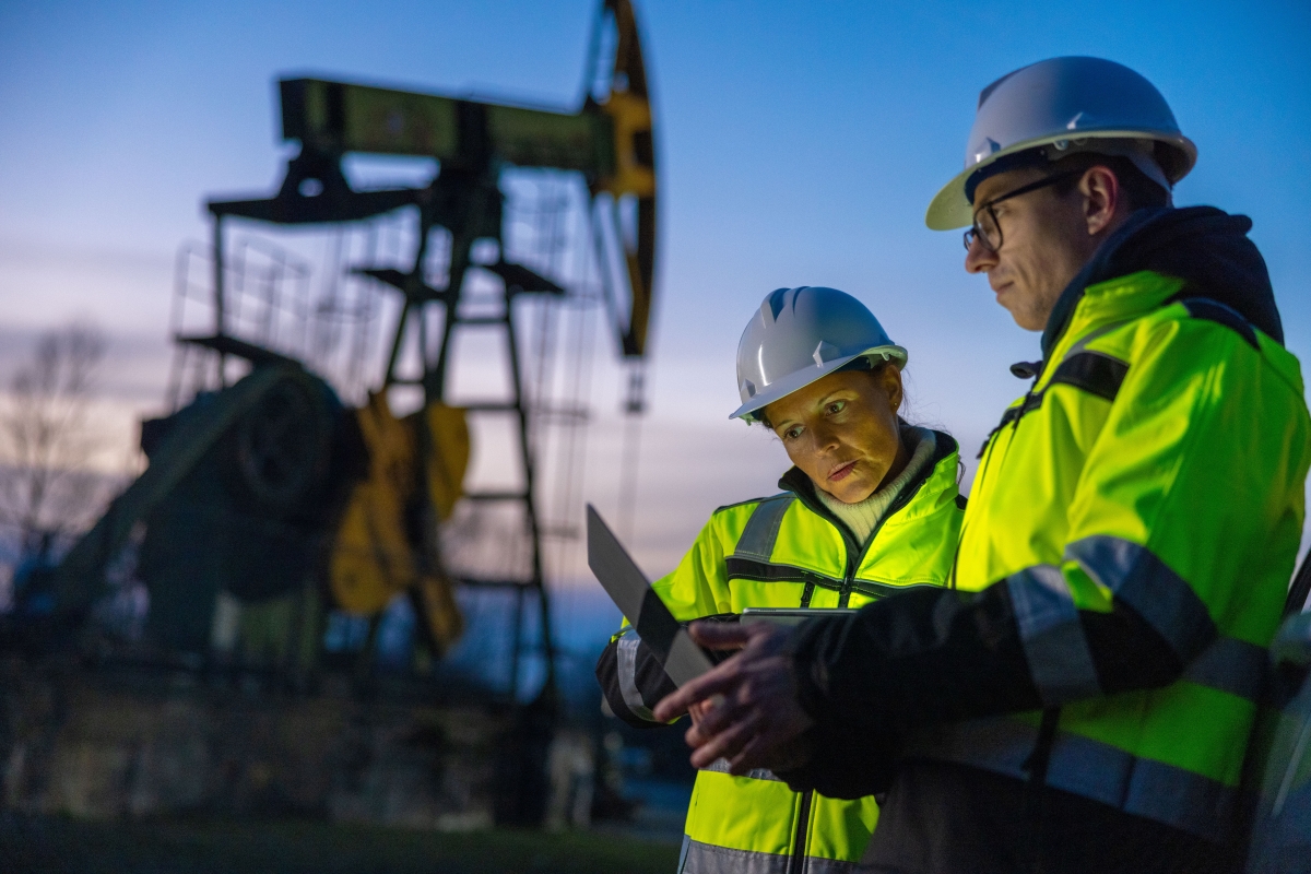 Male and female professionals in safety gear planning over laptop while standing close to oil pump during sunset.