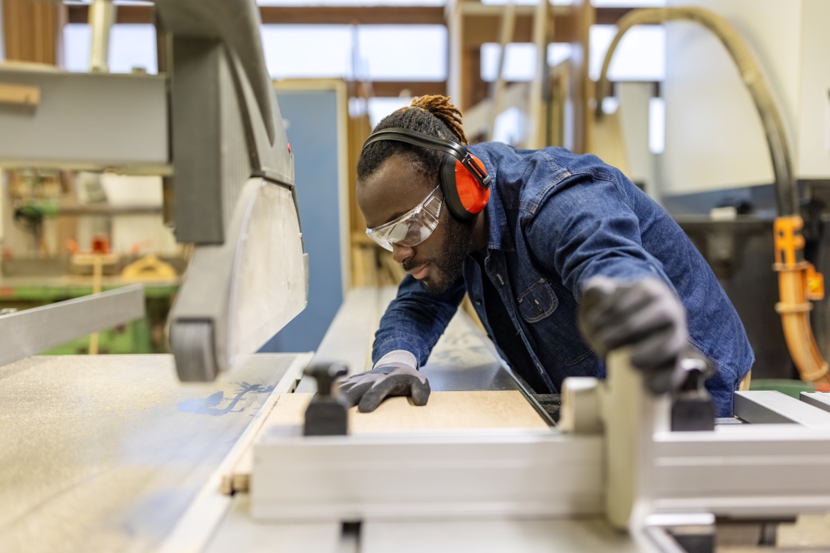 Black male carpenter wearing protective workwear cutting wood on electric circular saw in workshop.