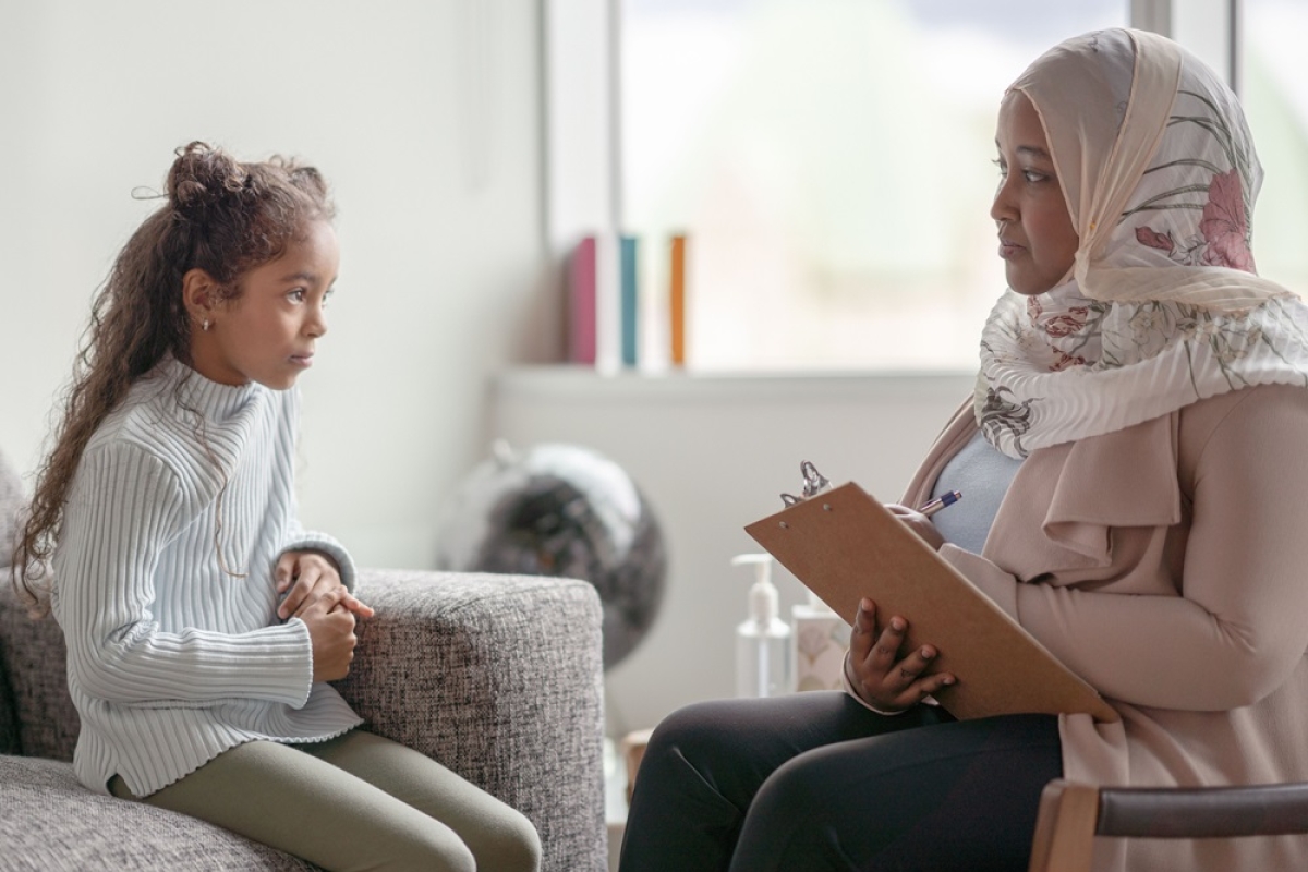 Muslim woman therapist wearing pink floral head scarf with clipboard talking with young female 