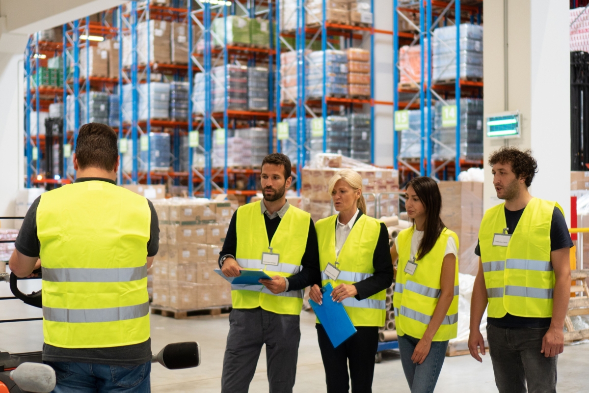 Group of peopl in a warehouse wearing safety vests listening to someone talk