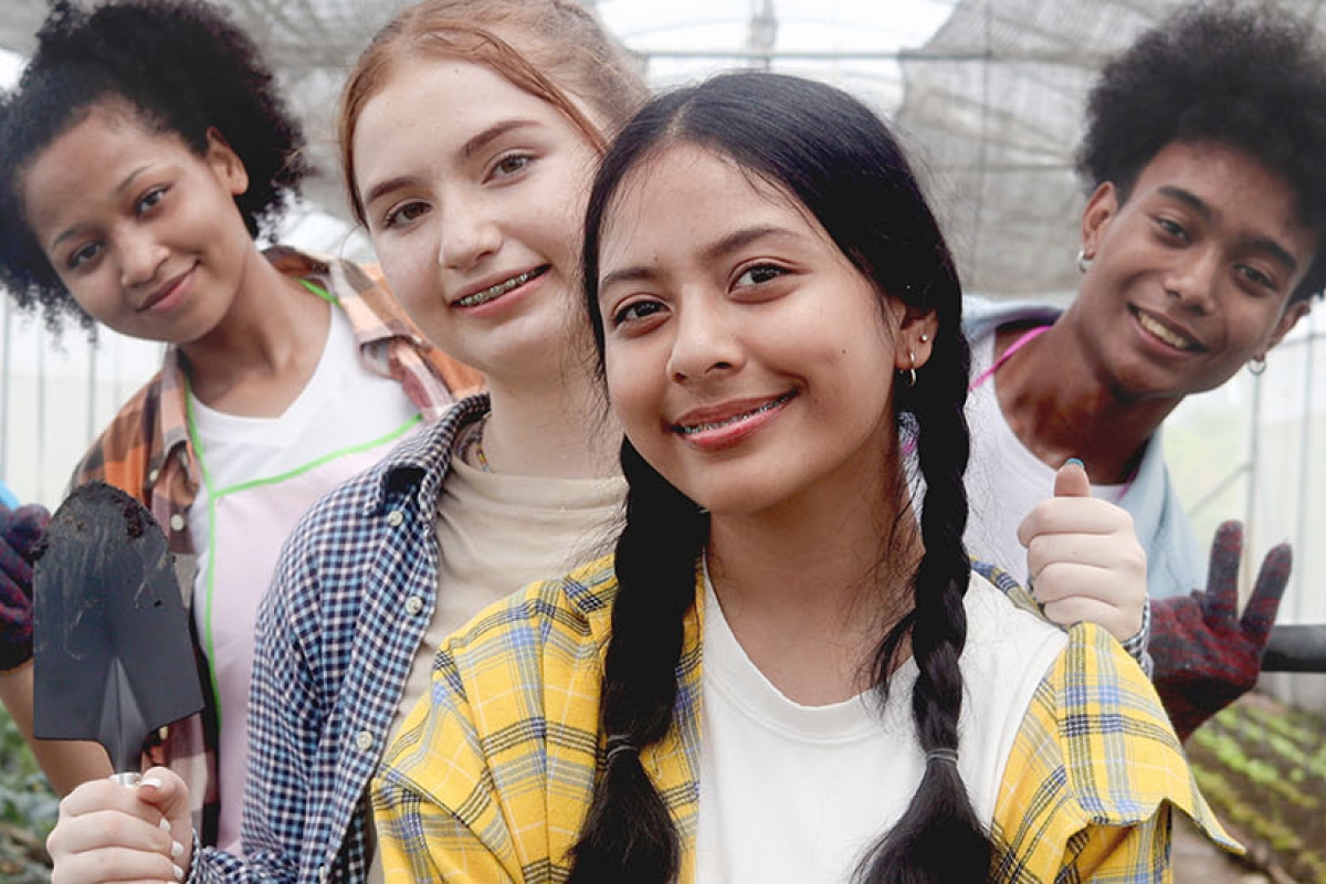 A diverse group of young people holding gardening tools and smiling in a commercial greenhouse