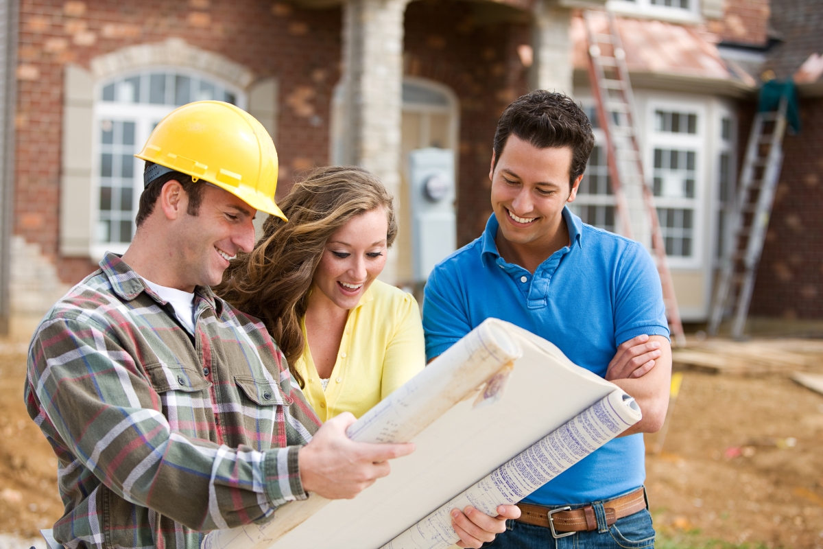 man in plaid shirt and yellow hard hat showing a man and woman project plans outside a house