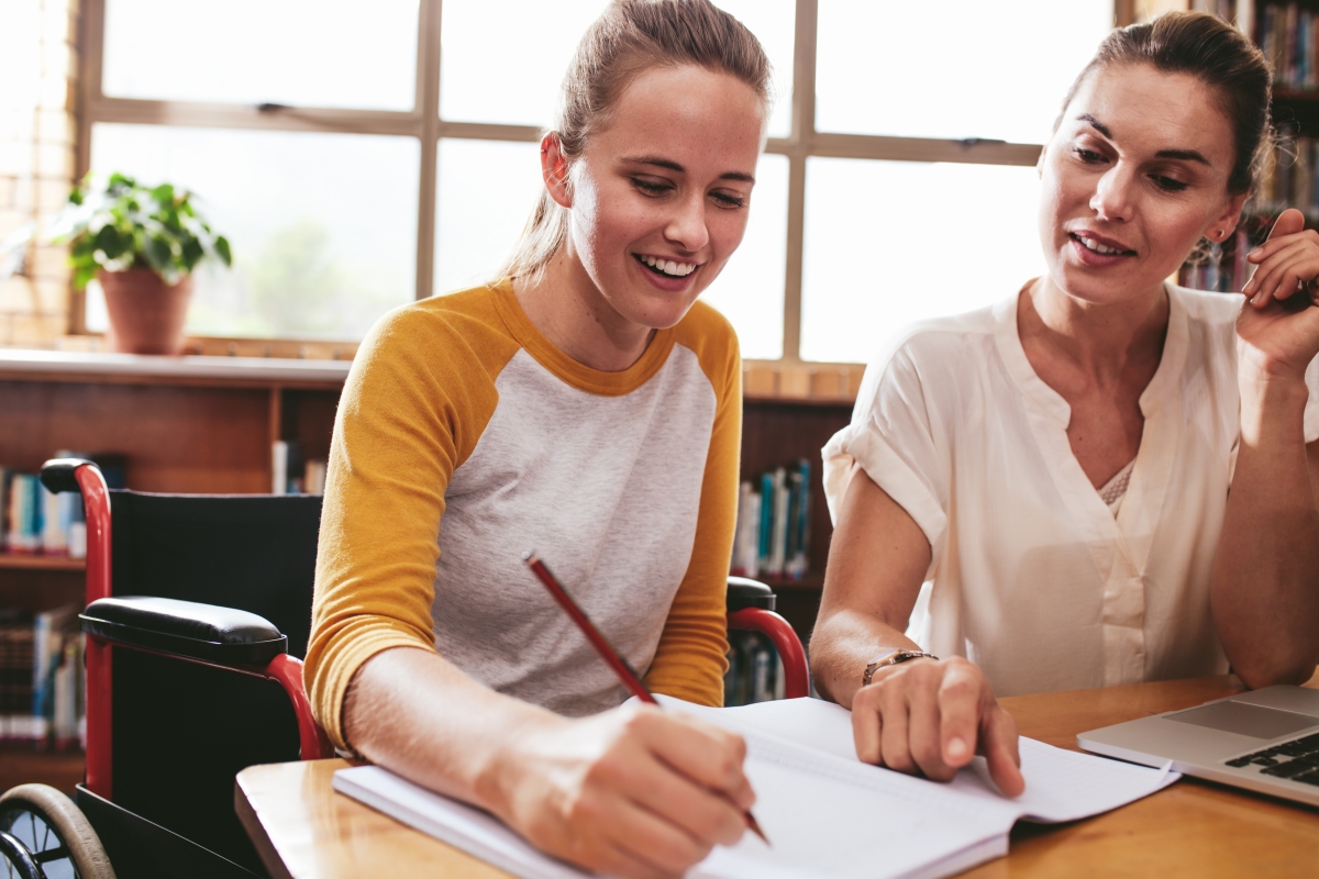 young woman writing in a notebook as another woman sitting next to her points at something on the page