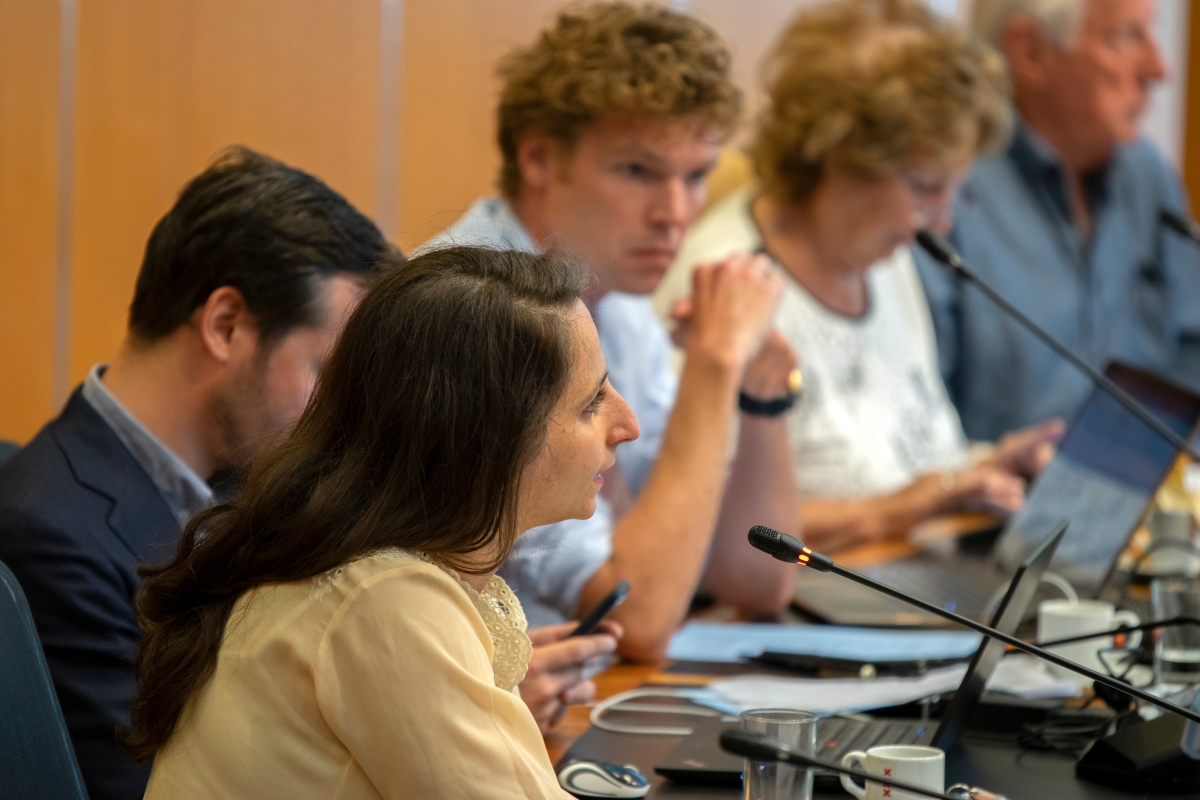 woman speaking into microphone while seated next to other people that also have microphones and laptops on the table in front of them