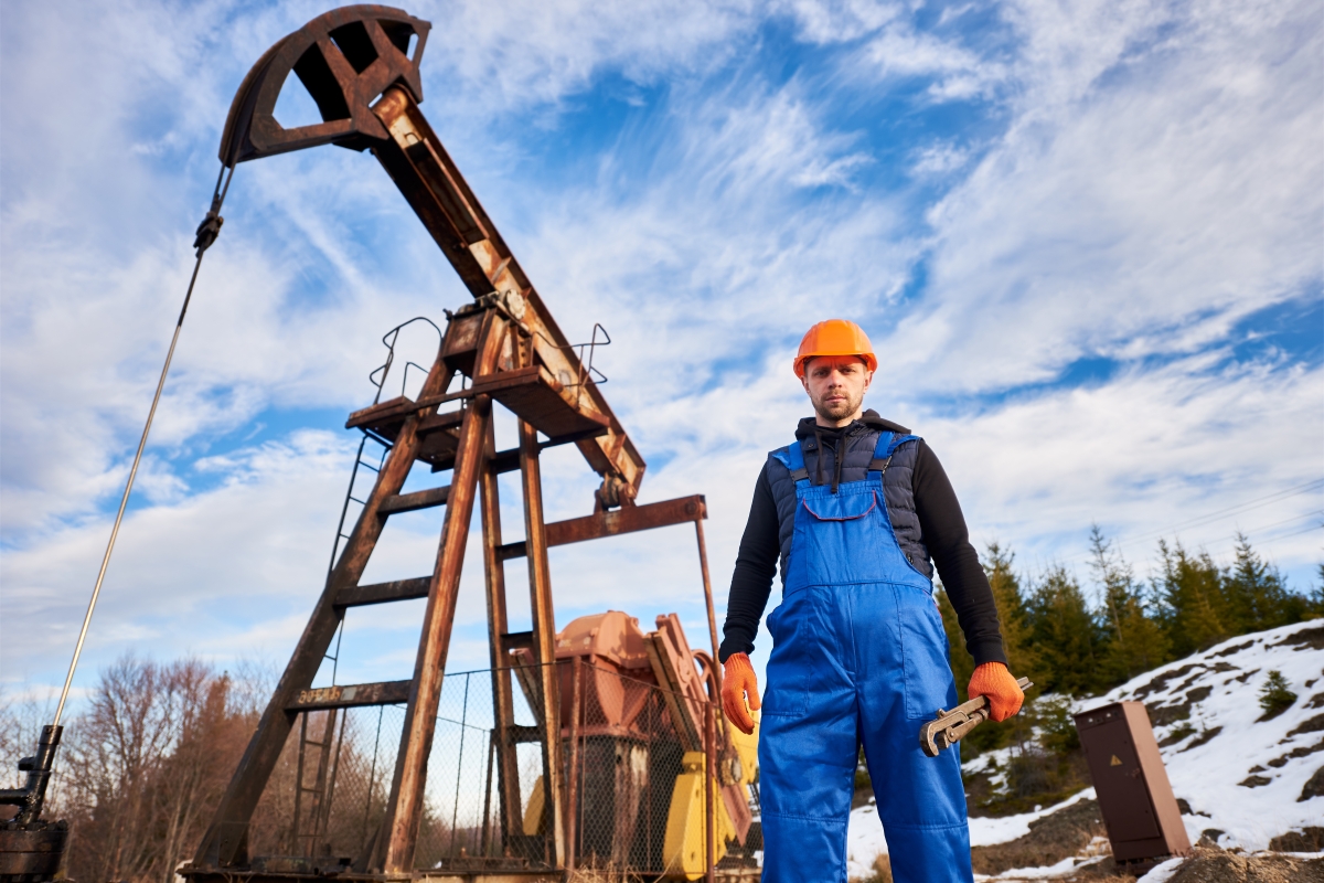 tall male worker in blue overalls holding a large tool and a oil well in the background